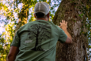 Man in a green Geelhout Tee touching the trunk of a tree