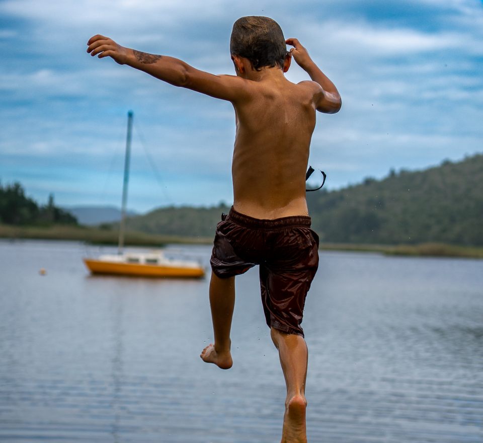 Boy leaping into a lake with a sailing boat behind